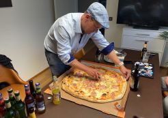 Riesige Pizza auf der MItte eines Tisches, umgeben von Getränken. Die Pizza wird eben vom Gastgeber mit blauer Mütze geschnitten. 